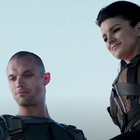 Gina Carano and Ed Skrein are standing next to each other, looking downwards.